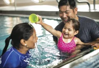 Father and daughter in a swim lesson. The daughter is playing with her instructor by pouring water on her head.