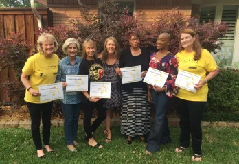 A group of seven women standing outdoors in front of a house and shrubs, smiling and holding certificates. Two women wear yellow “LIVESTRONG at the YMCA” shirts, while others are dressed casually. The group appears to be celebrating an achievement or program completion together.