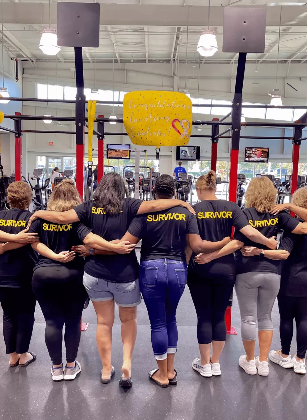 A group of people, all wearing black T-shirts with the word "SURVIVOR" printed in yellow on the back, stand together in a gym, facing away from the camera with their arms around each other.