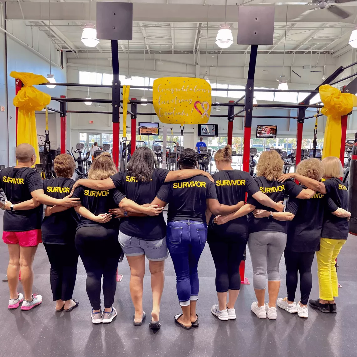 A group of people, all wearing black T-shirts with the word "SURVIVOR" printed in yellow on the back, stand together in a gym, facing away from the camera with their arms around each other. 