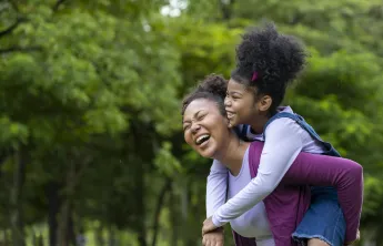 African American mother is playing piggyback riding with her young daughter while having a summer picnic in the public park