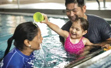 Father and daughter in a swim lesson. The daughter is playing with her instructor by pouring water on her head.