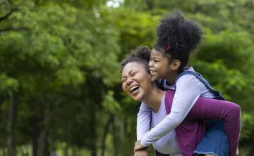 African American mother is playing piggyback riding with her young daughter while having a summer picnic in the public park
