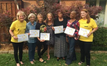 A group of seven women standing outdoors in front of a house and shrubs, smiling and holding certificates. Two women wear yellow “LIVESTRONG at the YMCA” shirts, while others are dressed casually. The group appears to be celebrating an achievement or program completion together.